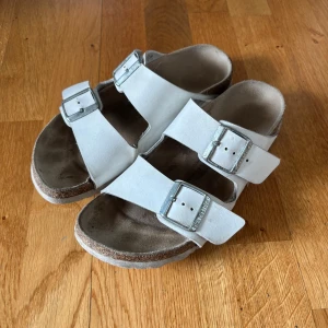 Vita sandaler från Birkenstock - Snygga vita sandaler från Birkenstock med två breda remmar och stora silverspännen. Skorna har en klassisk korksula och mjuk fotbädd för extra komfort. Perfekta för varma dagar och enkel att matcha med allt.