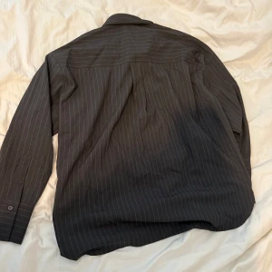 Ny Svart randig skjorta från NA-KD - Snygg svart skjorta från NA-KD med tunna vita ränder. Klassisk krage, knäppning framtill och långa ärmar med manschett. Skjortan har en rak och lite loose passform, perfekt till jeans eller dressade byxor. Materialet känns mjukt och följsamt.