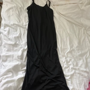 Svart långklänning med smala axelband - En stilren svart långklänning med smala justerbara axelband. Klänningen har en rak och enkel siluett, perfekt för dig som gillar minimalistisk stil. Materialet känns mjukt och följsamt mot kroppen. Passar dig som vill ha en klassisk och tidlös look.