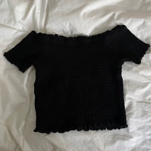 Svart offshoulder topp från Primark - En svart offshoulder topp från Primark med smockad struktur och volangkant vid halsringning och ärmslut. Toppen är kort och sitter tight, perfekt för dig som gillar en trendig och enkel stil. Materialet är mjukt och stretchigt för en bekväm passform.