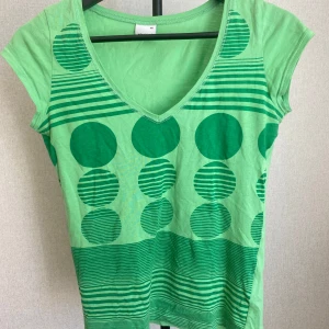 Grön t-shirt med unikt mönster! - Snygg t-shirt med v-ringning och korta ärmar. Framsidan har ett coolt grafiskt mönster med stora cirklar och ränder i olika gröna nyanser. Tillverkad i mjuk bomull, perfekt plagg för att liva upp outfiten!