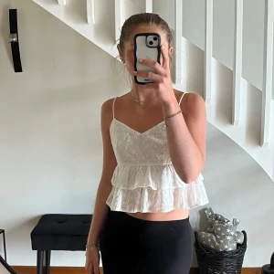 Volangtopp med broderi  - Superfin vit linnetopp med tunna axelband, v-ringning och dubbla volanger. Toppen har broderade små blommor, är något croppad och helt oanvänd. Perfekt för varma dagar och ger en söt, somrig vibe.