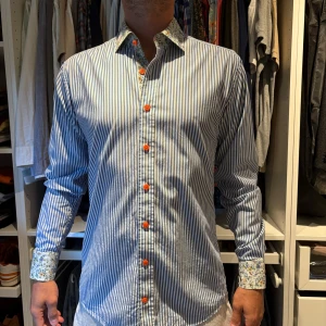 Randig skjorta med blommiga detaljer - Egen design från Tailorstore. Motsvarar storlek 39/M. Snygg skjorta med blå och vita ränder, orange knappar och blommigt tyg på insidan av krage och manschetter. Skjortan har lång ärm och klassisk krage, perfekt för dig som vill sticka ut lite extra.