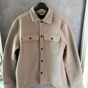 Beige overshirt med fickor och knappar - Stilren beige overshirt med klassisk krage och svarta knappar framtill. Två stora bröstfickor med lock ger en cool utility-vibe. Jackan är i ett mjukt ull-liknande material och har lång ärm. Perfekt för lager-på-lager och enkel att matcha.