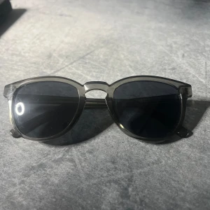 Svarta runda solglasögon i plast - Snygga svarta solglasögon med rundad form och transparent plastbåge. Glasen är mörka och ger en cool vibe, perfekt för att lyfta din stil. Passar dig som gillar klassisk design med en modern twist.