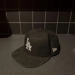 Svart LA-keps från New Era 7 3/8 - Snygg svart keps från New Era med LA-logga framtill i vitt. Klassisk 59FIFTY-modell med böjd skärm och broderade detaljer. Tillverkad i polyester och bomull, perfekt för dig som gillar streetwear och sportig stil.