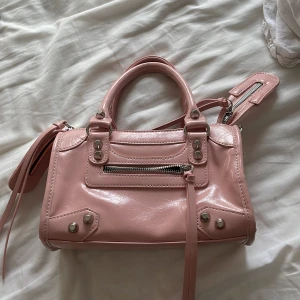 Rosa handväska med dragkedja - Söt rosa handväska i glansigt skinnimitation med silverfärgade detaljer och dragkedjor. Väskan har både handtag och en lång axelrem, samt dekorativa nitar framtill. Perfekt storlek för det viktigaste och snygg att bära till vardagslooken.