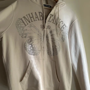 Beige hoodie med fjäril från BBL - Snygg beige hoodie från: By Ben Livermore med rhinestone fjärilsmotiv och texten 'INHABITANCE' framtill. Tröjan har huva, snörning, fickor och dragkedja. 