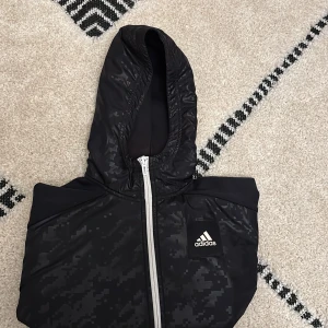 Svart vindjacka från Adidas med huva - Svart vindjacka från Adidas med coolt pixelmönster och vit dragkedja. Jackan har huva och Adidas-logga på bröstet. Perfekt för dig som gillar sportig stil och vill ha något lätt och smidigt till vardagen.