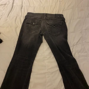 Svarta jeans från True Religion - Säljer ett par svarta jeans från True Religion med slitningar och trasiga detaljer på benen. Jeansen har klassiska bakfickor med broderade sömmar och en bootcut passform. Perfekta för dig som gillar en edgy och avslappnad stil.