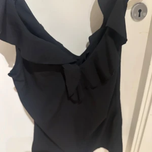 Svart ärmlös volangtopp - Snygg svart ärmlös topp med volangdetalj längs halsringningen. Toppen har en djup v-ringning och en feminin, flowy känsla. Från vero moda 