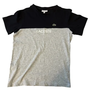 Grå och svart t-shirt från Lacoste - Snygg t-shirt från Lacoste med svart överdel och grå nederdel. Klassisk rund hals och korta ärmar. Framsidan har Lacoste-logga och broderad krokodil på bröstet. Tillverkad i mjuk bomull som är skön mot huden. Perfekt för en chill och stilren look.