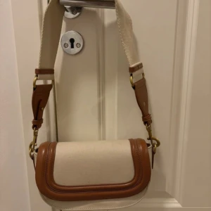 Beige och brun axelväska med bred rem - Snygg axelväska i beige canvas med bruna skinn-detaljer och bred. Väskan har guldfärgade metalldelar och en stilren. Perfekt för dig som vill ha en trendig och praktisk väska  . Köptes för 1 år sedan och använt 3/4 gånger. 