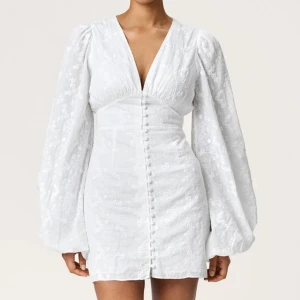 Vit kort klänning med puffärm - Supersnygg vit kort klänning med v-ringning och puffiga långärmar. Klänningen har broderade blommor, knappar framtill och markerad midja. Perfekt för dig som vill sticka ut med en romantisk och trendig look. Den saknar dock en knapp men det finns i för att sätta tillbaka. 