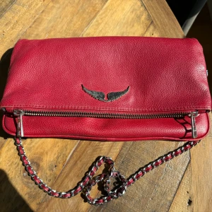 Röd skinn axelväska från Zadig & Voltaire - Snygg röd axelväska i skinn från Zadig & Voltaire med silverfärgad kedja och coolt vingemblem framtill. Väskan har dragkedja längs kanten och en mjuk, pebble-textur. Perfekt storlek för det viktigaste och ger en edgy vibe till din outfit. ÄKTA ZADIG & VOLTAIRE köptes för 4999 säljes nu för 3000 kr, men priset är diskuterbart 
