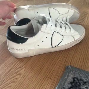Philippe Model vita sneakers strl 39 - Snygga vita Philippe Model sneakers med svart detalj på hälen och grå mocka vid tån. Klassisk låg modell med rund tå och platt sula. Skorna har snörning och är tillverkade i skinn och mocka, med coola kontrastdetaljer och logga på baksidan. Nypris 3299kr