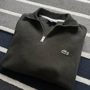 Lacoste Tröja/ Halvzip - Säljer en stilren olivgrön tröja från Lacoste med dragkedja och klassisk krokodillogga på bröstet. Perfekt för en avslappnad men snygg look. Passar bra till både jeans och chinos.