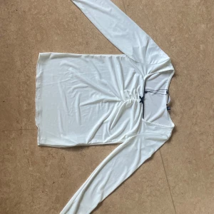 Vit långärmad topp med rosett från vero moda - Supersöt vit långärmad topp från vero moda med snygg rynkning framtill och en liten svart rosett vid halsen. Materialet är mjukt och har en lätt glansig finish, vilket ger en lyxig känsla. Svart kantdetalj runt halsen och smal passform. Perfekt för att lyfta din outfit.