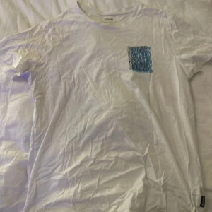 Vit t-shirt med färggrafiskt tryck - Säljer en vit t-shirt från Yourturn med coolt grafiskt tryck i blått och rött på ryggen och mindre tryck på bröstet. T-shirten har rund hals och korta ärmar, perfekt för dig som gillar streetstyle och vill sticka ut lite extra.