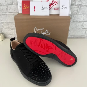 Christian Louboutin svarta sneakers med nitar - Säljer ett par svarta sneakers från Christian Louboutin med ikonisk röd sula och coola svarta nitar på tån. Skorna har snörning och är tillverkade i skinn med en platt sula. Perfekt för dig som vill sticka ut med en edgy och lyxig stil.
