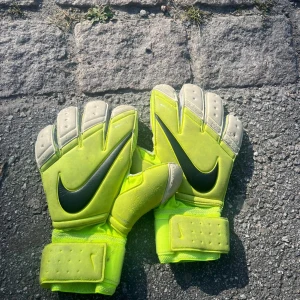 Nike målvaktshandskar i neon - Säljer ett par grymma målvaktshandskar från Nike i neon-gult med svarta swoosh-loggor. Handskarna har breda kardborreband runt handleden och insidan är ljusgrå med extra grepp. Perfekt för dig som vill sticka ut på fotbollsplanen! Storlek 9,5. Se bilder för skick och storlek.Dyra när dem köptes!