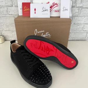 Christian Louboutin svarta sneakers med nitar - Säljer ett par svarta sneakers från Christian Louboutin med ikonisk röd sula och coola svarta nitar på tån. Skorna har snörning och är tillverkade i skinn och mocka för en lyxig vibe. Perfekta för dig som vill sticka ut med exklusiv design och edgy detaljer.
