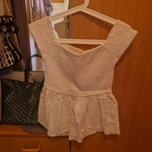 Vit offshoulder topp från Hollister - Supersöt vit offshoulder topp från Hollister med smockad överdel och volangig spets nertill. Toppen har en romantisk vibe med sin struktur och breda axelband. Perfekt för dig som gillar en feminin och somrig stil!