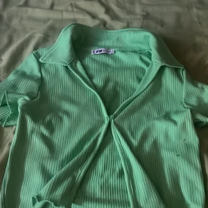 Ribbad grön blus från FB Sister - Snygg ribbad blus i ljusgrönt från FB Sister. Modellen har korta ärmar, krage och knäppning framtill med en knapp. Materialet är stretchigt och ger en fräsch vibe, perfekt för dig som gillar färg och trendiga detaljer.