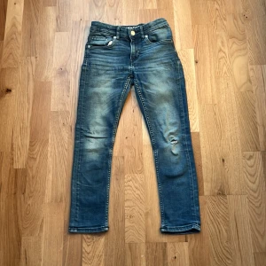 Blå skinny jeans från &Denim, super stretch - Säljer ett par blå skinny jeans från &Denim med super stretch. Jeansen har slitningar och hål på ena benet för en cool look. Klassisk femficksmodell och smal passform. Perfekta för dig som gillar bekväma och trendiga jeans.