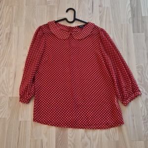 Röd prickig blus med krage från Koton - Säljer en röd blus från Kotonl med vita prickar och rund krage. Blusen har trekvartsärm och stängs med dragkedja i ryggen. Materialet är lätt och luftigt, perfekt för dig som gillar retrovibbar och vill sticka ut med ett gulligt mönster.
