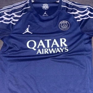 PSG blå fotbollströja Jordan - Snygg marinblå PSG fotbollströja från Jordan med vita detaljer och loggor. Tröjan har korta ärmar, rund hals och coola vita mönster på axlarna. Materialet är lätt och andas, perfekt för träning eller match. Qatar Airways tryck på bröstet.