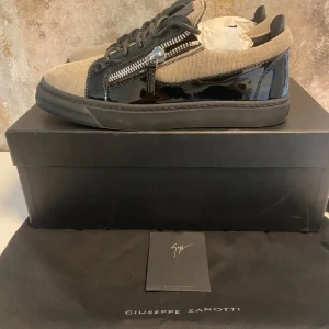 Giuseppe Zanotti sneakers med dragkedja - Snygga sneakers från Giuseppe Zanotti i beige textil och svart lackdetalj. Skorna har svarta snören, dragkedjor på sidorna och en robust svart sula. Perfekt för dig som gillar exklusiv och modern streetstyle.