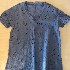 Blå blus med broderade blommor - Supersöt blå blus med v-ringning och volangkant. Blusen har korta ärmar med resår och är täckt av broderade blommor som ger en romantisk vibe. Perfekt för dig som gillar detaljer och en luftig känsla. Tillverkad i bomull för extra komfort.