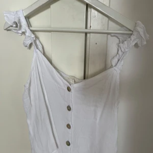 Vit linnetopp med knappar - Superfin vit linnetopp med volangiga axelband och dekorativa knappar framtill. Toppen är ärmlös och har en luftig, lätt passform som passar perfekt till varma dagar. Snygg att styla med jeans eller kjol för en fräsch look.