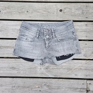 Låga jeansshorts  - Jeansshorts i grå denim med råa kanter och dubbla knappar. Sitter supersnyggt med låg midja och kort passform – perfekt till sommarens alla looks. Helt nya och aldrig använda, stretchiga och sköna i materialet. Midjemått 36cm rakt över och 78cm runt om hela!
