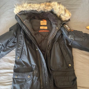 Svart Parajumpers parkajacka med päls - Säljer en svart Parajumpers parkajacka i storlek small med snygg päls på huvan och flera praktiska fickor. Jackan har coola detaljer som patch på ärmen, dragkedja och knappar. Perfekt för kalla vinterdagar och har en riktigt streetig vibe.