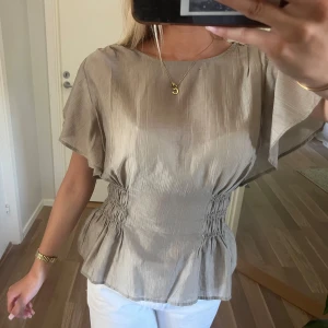 Beige blus med smock i midjan - Säljer en snygg ljusgrön blus med korta, vida ärmar och smockad midja som ger en markerad siluett. Blusen har lätt glansigt tyg och en avslappnad passform med volang nertill. Perfekt för dig som gillar stilrena och trendiga plagg.