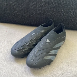 Adidas Predator fotbollsskor svarta Pris kan diskuteras  - Säljer ett par svarta Adidas Predator fotbollsskor med silvriga detaljer och klassiska ränder på sidorna. Skorna har en strumpliknande passform utan snörning och är tillverkade i syntetmaterial för bästa bollkontroll. Perfekta för dig som vill dominera på planen. Änvänt i cirka 6 månader. Storlek 42 2/3