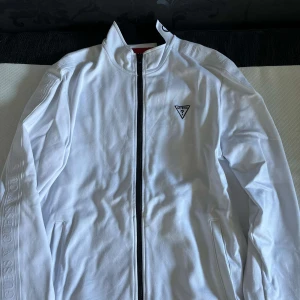 Vit zip från Guess med logga - Snygg vit vindjacka från Guess med svart dragkedja och broderad logga på bröstet. Jackan har hög krage med Guess-tryck och diskreta loggor längs ärmarna. Perfekt för dig som gillar stilrena och sportiga plagg. Köpt från guess i Los Angeles för 1000kr