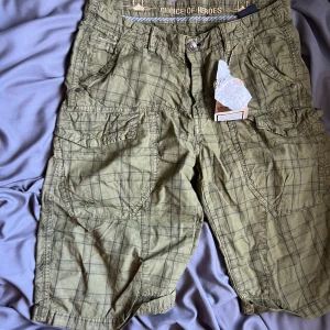 Gröna rutiga cargoshorts Alpha Industries - Snygga cargoshorts från Alpha Industries i olivgrönt med rutigt mönster. Shortsen har flera stora fickor med lock och knapp, samt klassisk cargostil. Perfekta för sommaren och riktigt sköna att bära. Stängs med knapp och dragkedja.