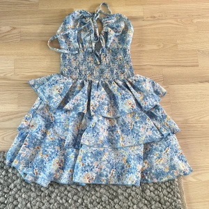 Blommig blå kortklänning med volanger - Supersöt kortklänning i blått med blommigt mönster och volanger. Klänningen har omlottknytning upptill, smockad midja och axelbandslös design. Perfekt för sommarens alla tillfällen, med luftig känsla och härliga färger.