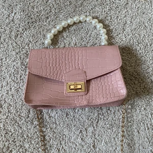 Rosa handväska med pärlhandtag - Säljer en ljusrosa handväska i syntet med croco-mönster, pärlhandtag och guldfärgad kedja. Väskan har ett snyggt vridlås i guld och stilren, boxig form. Perfekt accessoar för att lyfta din outfit.