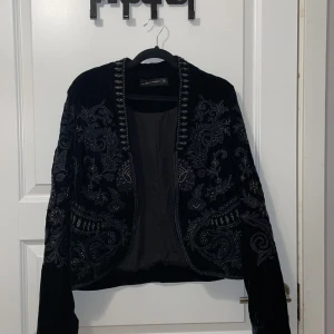 Svart broderad kavaj från Zara Woman - Säljer en svart kavaj från Zara Woman med snygga broderade detaljer i sammet. Jackan har lång ärm, öppen front och vackra mönster över hela plagget. Perfekt för dig som vill sticka ut med en unik och elegant look.