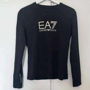 Svart långärmad EA7 Emporio Armani tröja - Svart långärmad tröja från EA7 Emporio Armani med silverfärgad logga på bröstet och ärmen. Slimmad passform och rund halsringning. Tillverkad i mjuk bomull som känns skön mot huden. Perfekt för dig som gillar stilrena och exklusiva plagg. storlek S