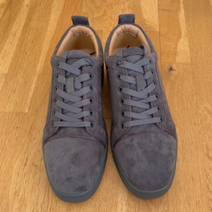 Grå mockasneakers från Christian Louboutin - Snygga sneakers i grå mocka från Christian Louboutin med ikonisk röd sula. Skorna har rund tå, platt sula och grå snörning. Insidan är fodrad i beige läder för extra komfort. Perfekta för dig som vill sticka ut med exklusiva detaljer.