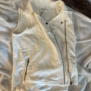 Vit väst från American Vintage - Snygg vit väst från American Vintage i modellen Patterson. Väst med klassisk krage, sned dragkedja framtill och dragkedjeficka. Tillverkad i mjukt bomullstyg, perfekt för lager-på-lager och stilrena outfits.