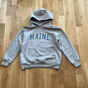 Beige hoodie MAINE från 157 - Snygg beige hoodie från 157 med MAINE-tryck i blått och vitt på bröstet. Tröjan har huva, ribbade muddar och en stor magficka framtill. Perfekt för chill dagar och enkel att matcha med jeans eller mjukisbyxor.