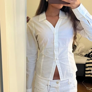 Figursydd skjorta - Säljer en vit skjorta med klassisk krage och knappar framtill. Skjortan har lång ärm och är figursydd med en liten slits nedtill framtill. Perfekt för dig som gillar stilrena och enkla plagg.