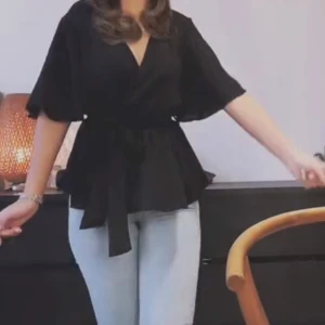 Svart omlottblus med knytband - Snygg svart omlottblus med v-ringning och vida trekvartsärmar. Blusen har ett knytband i midjan som ger en cool siluett och volang nedtill. Perfekt att styla med jeans eller kjol för en trendig look.