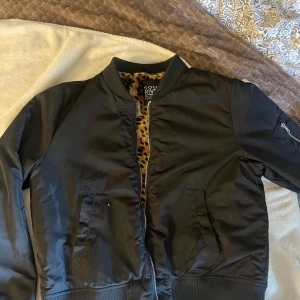 Svart bomberjacka med leopardfoder - Svart bomberjacka från Soul River i storlek M. Jackan har ett mjukt, leopardmönstrat foder och klassisk ribbad krage, muddar och nederkant. Utrustad med dragkedja framtill och fickor på sidorna samt ärmficka med dragkedja. Perfekt statement-plagg för din garderob.